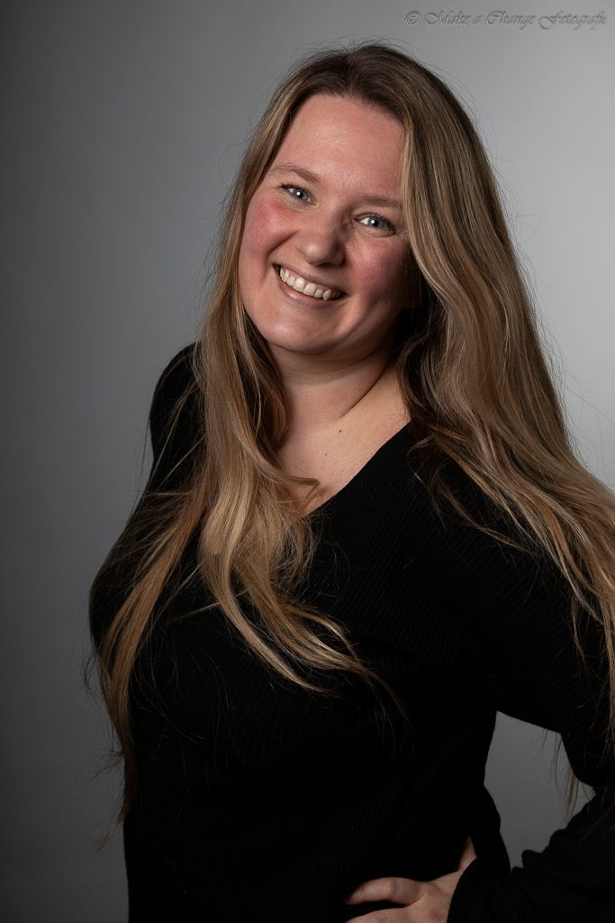 Een vrouw met lang, golvend blond haar, gekleed in een zwarte top, lacht en poseert tegen een grijze achtergrond.
