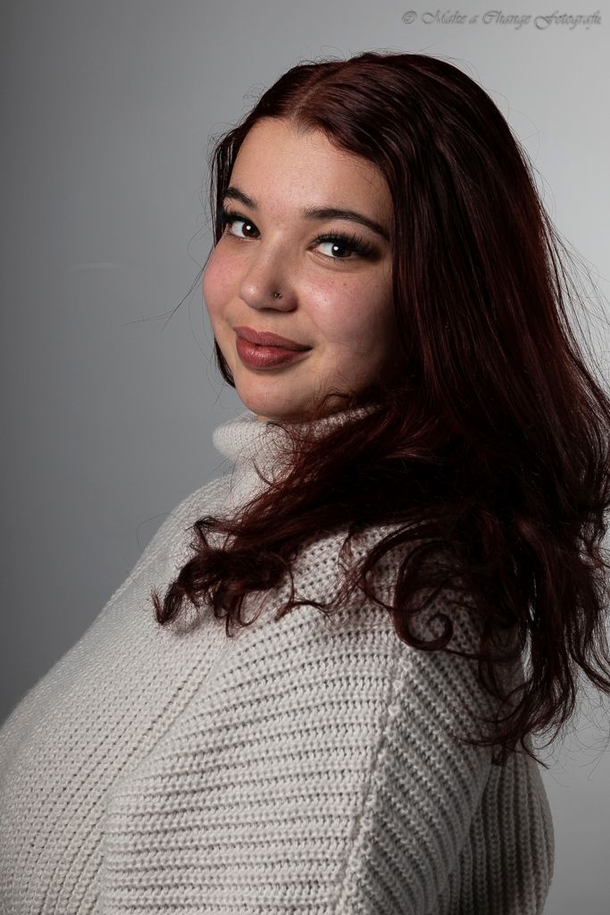 Vrouw met kastanjebruin haar in een crèmekleurige trui glimlacht over haar schouder tegen een grijze achtergrond.