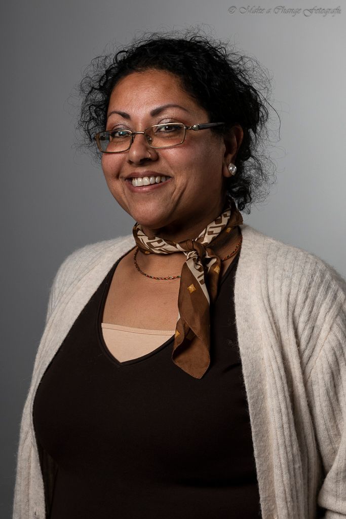 Vrouw met bril en sjaal, lachend, poseert voor een grijze achtergrond.