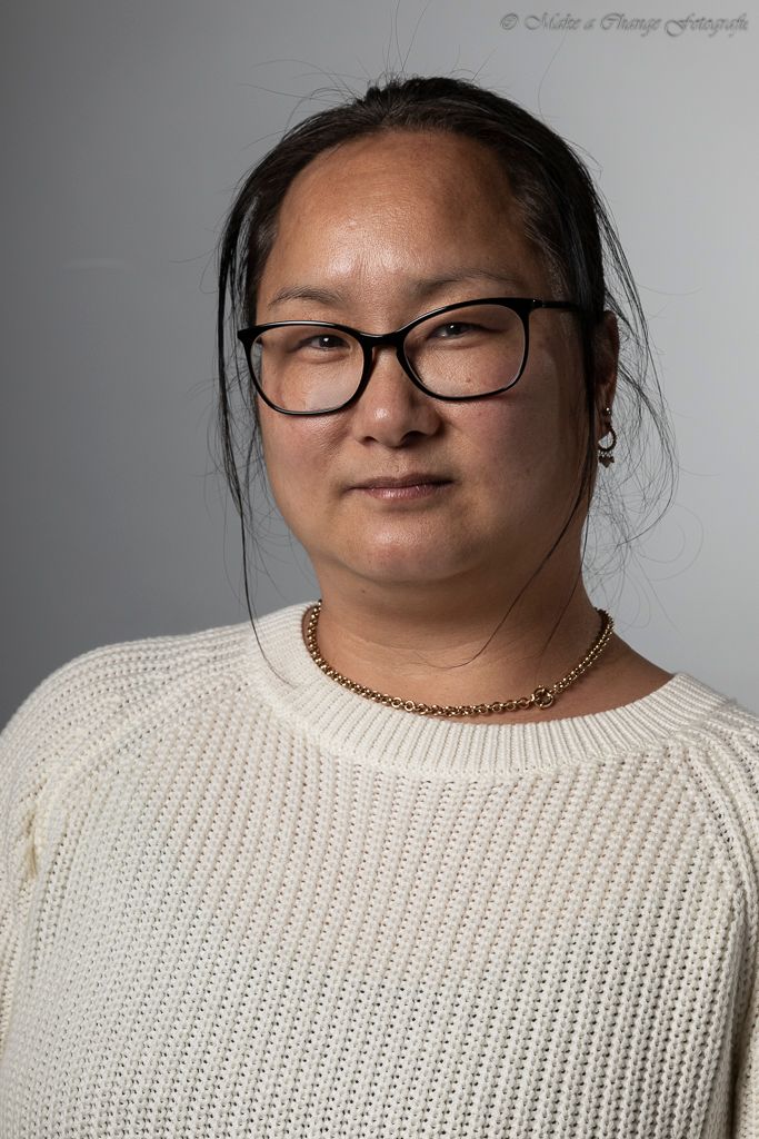 Vrouw met bril en gouden ketting, gekleed in een witte trui, kijkt in de camera.