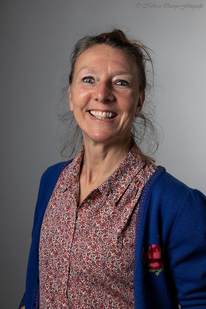 Een lachende vrouw, gekleed in een blauw vest en een blouse met bloemenprint, staat tegen een grijze achtergrond.