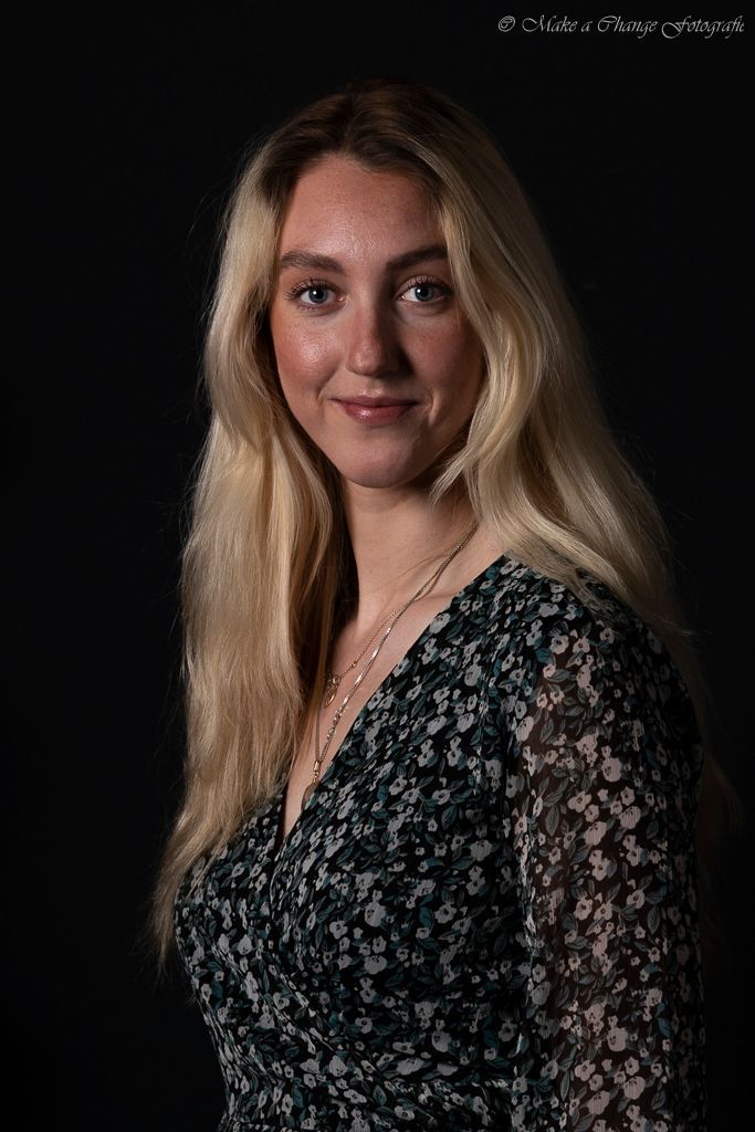Vrouw met lang blond haar, lachend, in een bloemenjurk, tegen een donkere achtergrond.