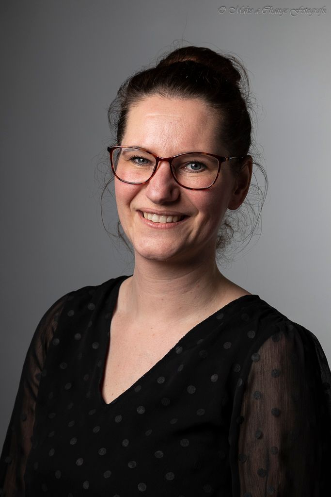 Vrouw met bril lacht, knotkapsel, grijze achtergrond.