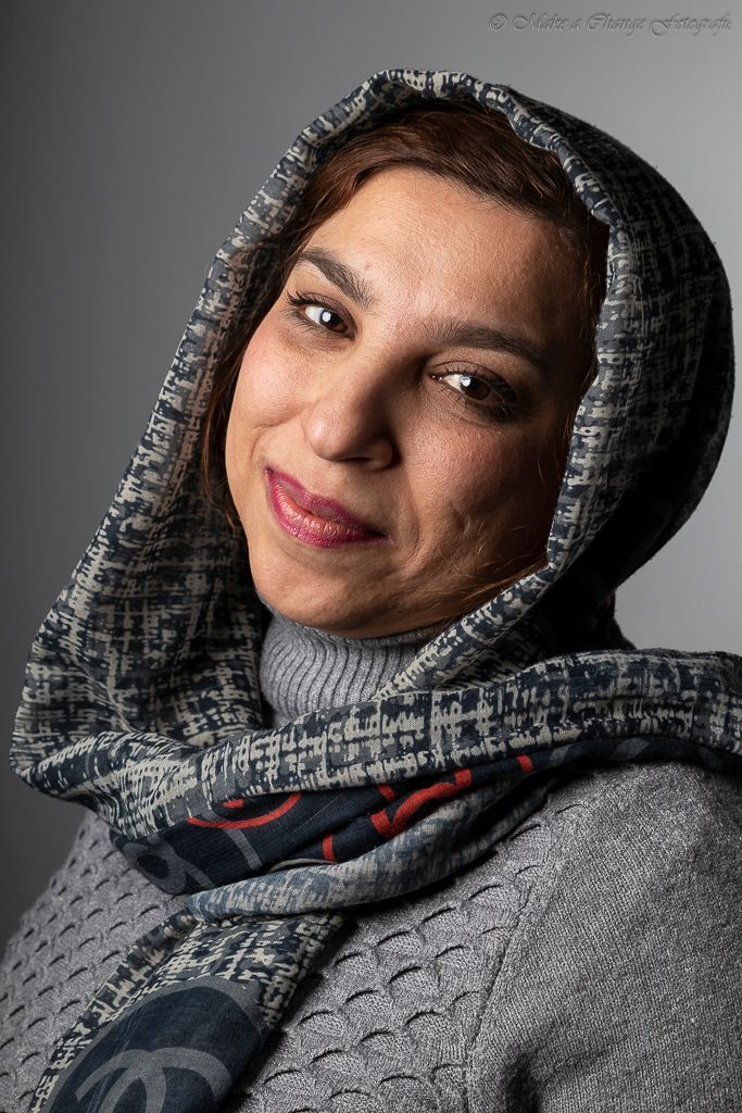 Vrouw met een hoofddoek met patroon en een grijze trui, lachend tegen een neutrale achtergrond.