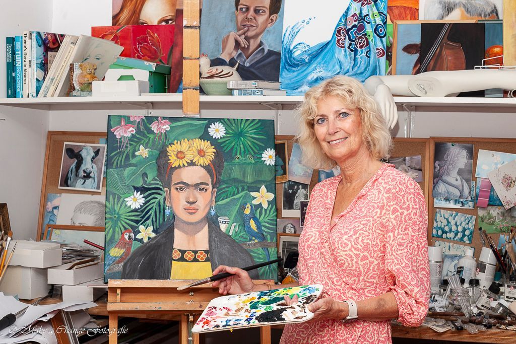 Vrouw met palet en penseel, glimlachend naast een schilderij van Frida Kahlo in een kunststudio.