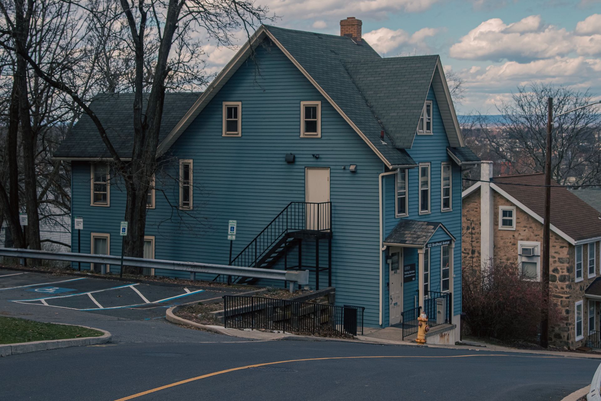 Blue house on a sloped street at dusk, with leafless trees and neighboring homes nearby.