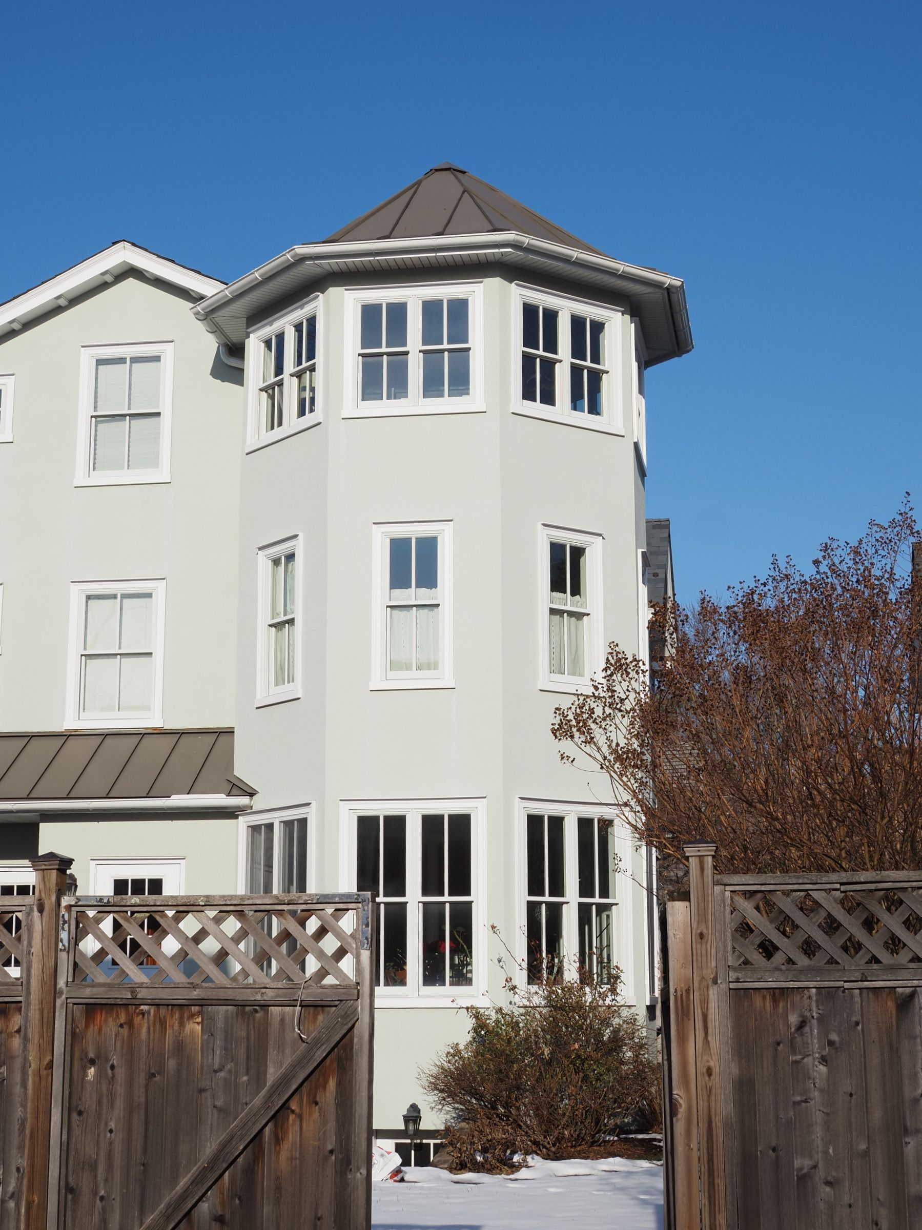 White multi-story house with bay windows, brown roof, and wooden fence under a blue sky