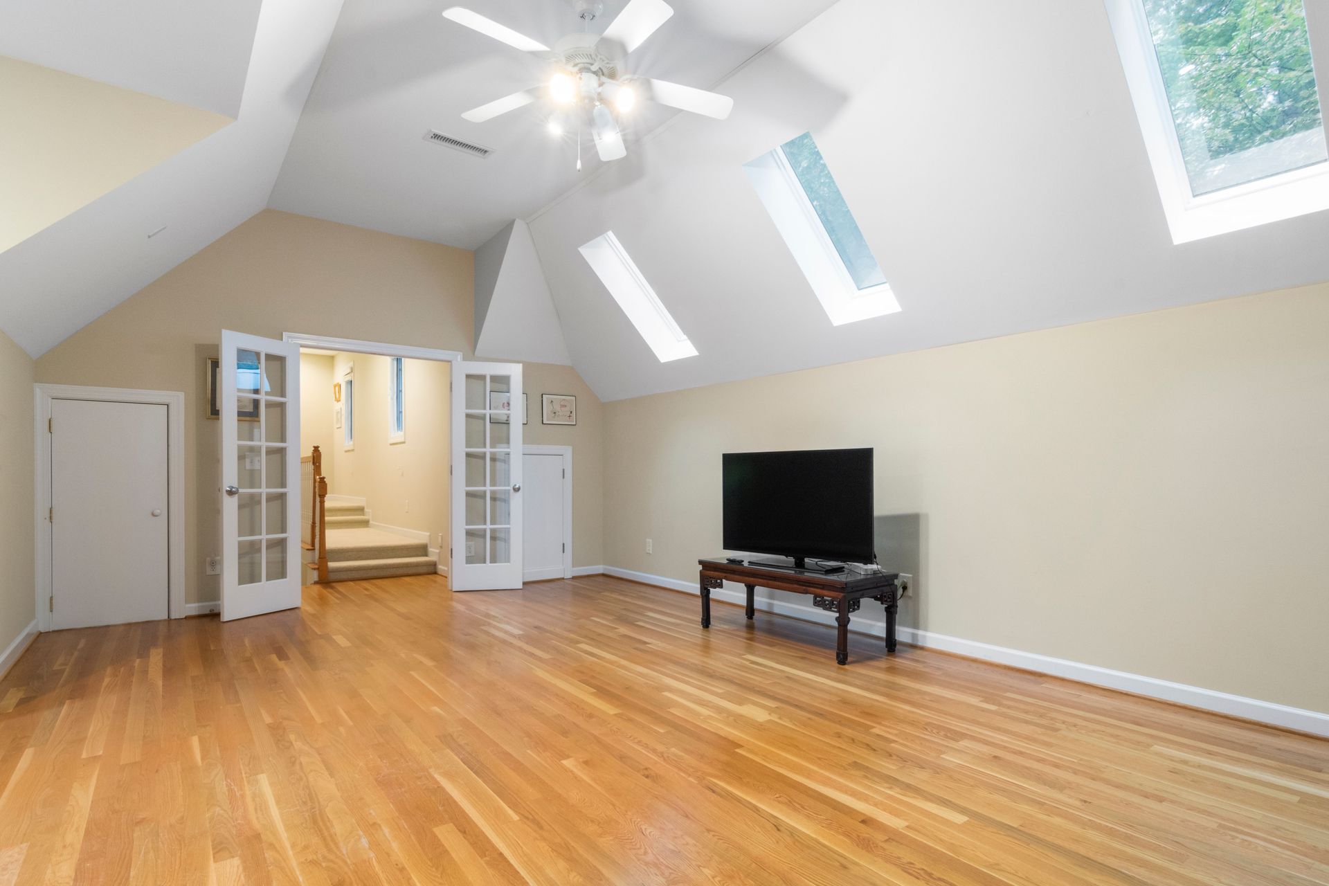 Bright, empty attic room with hardwood floors, skylights, ceiling fan, and a TV on a low table.