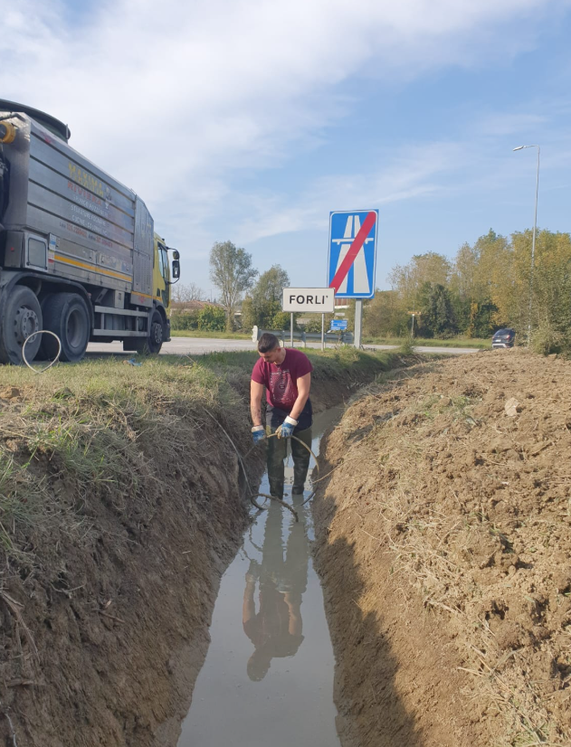 Un operaio in una trincea ispeziona l'acqua fangosa a lato di una strada, con un grosso camion e un cartello stradale nelle vicinanze.