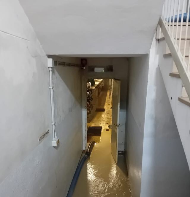 Corridoio del seminterrato allagato con acqua stagnante e scale che conducono al piano superiore sulla destra.