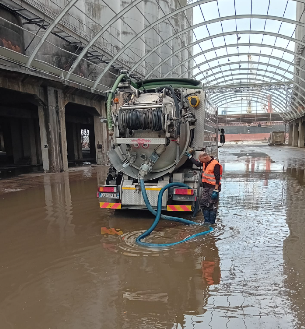 Un camion della nettezza urbana pompa acqua alluvionale all'interno di un edificio industriale coperto, con operai che indossano giubbotti arancioni.
