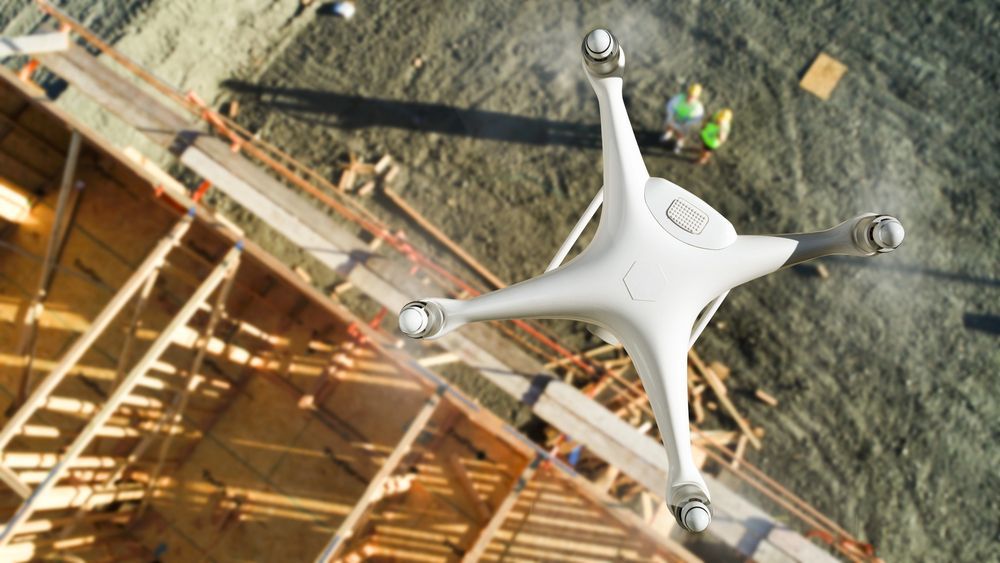 Un drone quadricottero bianco vola sopra un cantiere edile con struttura in legno e operai sottostanti.