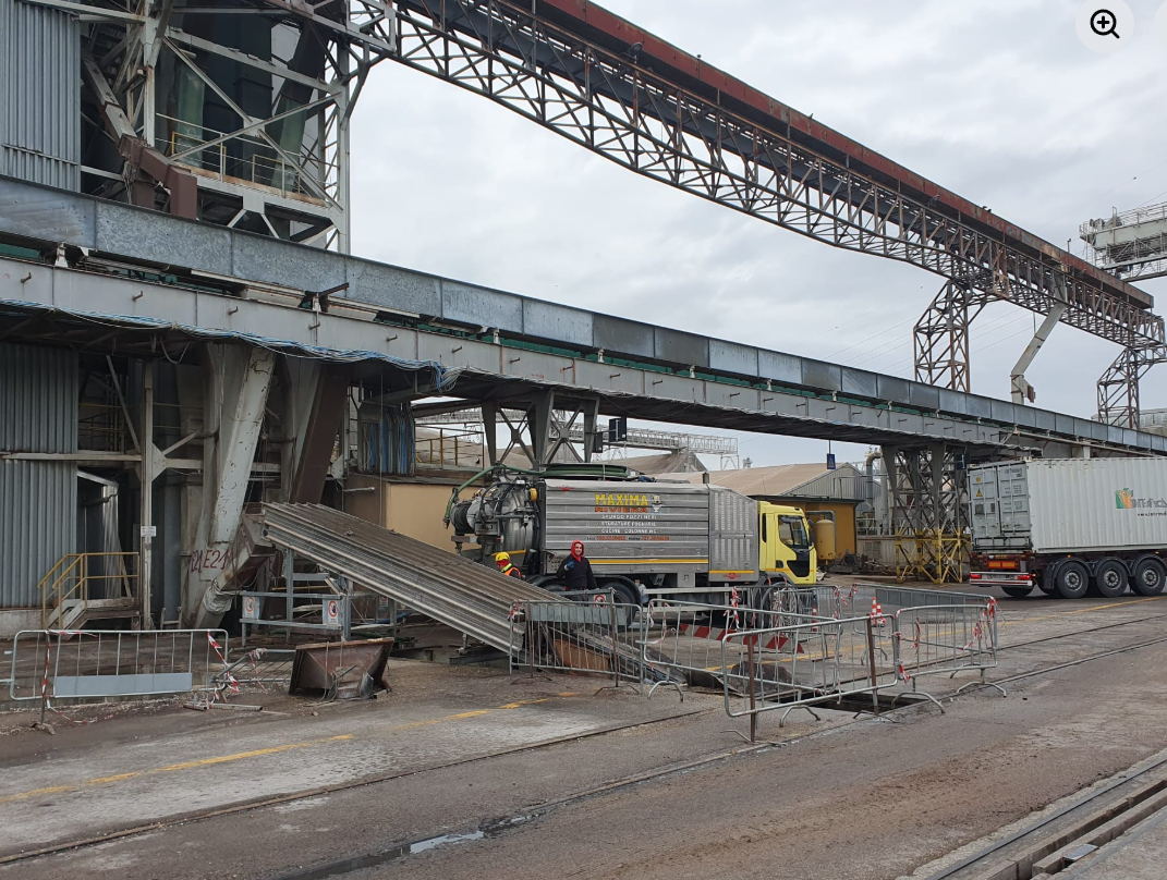 Impianto industriale con nastri trasportatori, un camion giallo e vagoni ferroviari sotto un cielo nuvoloso.