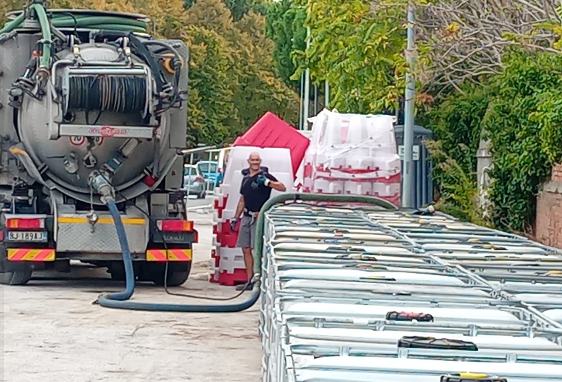 Un camion della nettezza urbana con tubi flessibili pulisce una fila di grandi serbatoi metallici in un'area industriale.