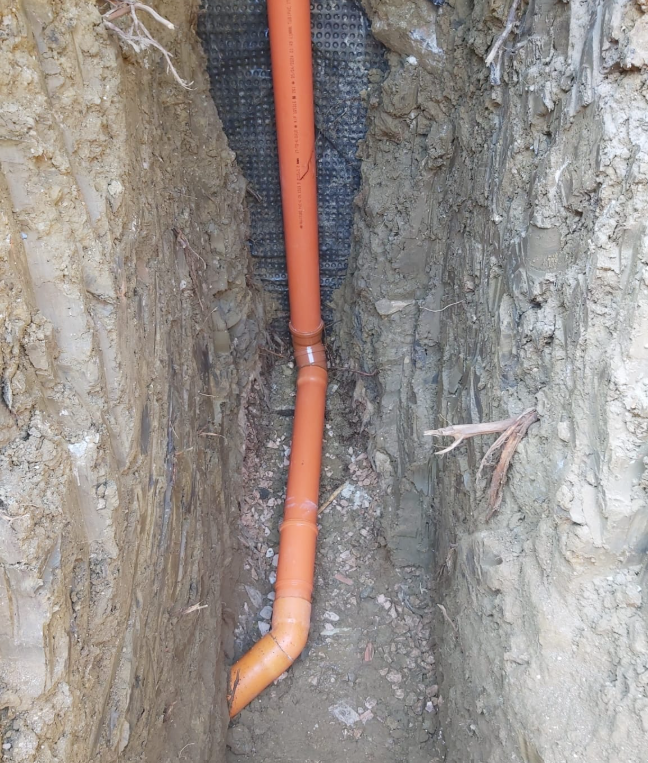 Tubo di drenaggio arancione che corre verticalmente in una stretta trincea scavata nella terra e nella roccia.