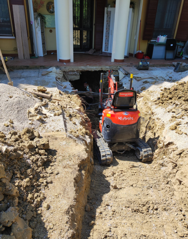 Un mini escavatore rosso in una profonda trincea accanto a una casa in costruzione.