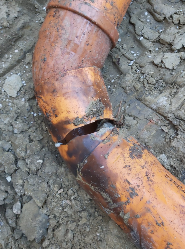 Giunto di tubo arancione arrugginito con una sezione incrinata e che perde, su terreno ghiaioso.