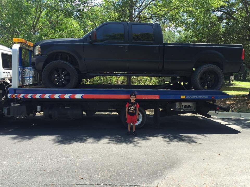 Black pickup truck on a flatbed tow truck, with a small dog standing underneath in a parking lot.