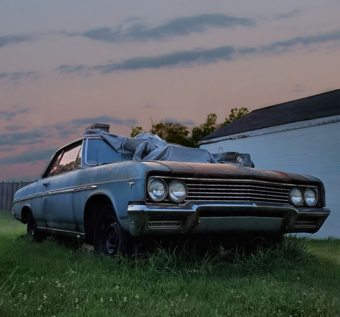 Abandoned blue vintage car beside a white building at dusk, with overgrown grass and a pink sky