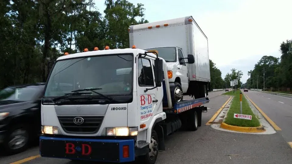 Tow truck carrying a white box truck on a road beside a grassy median and trees