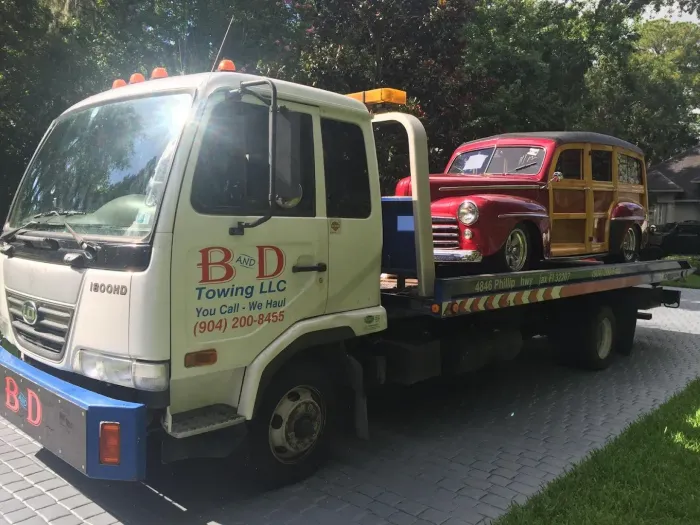 Tow truck carrying a red vintage car on a flatbed, parked on a sunlit road beside trees