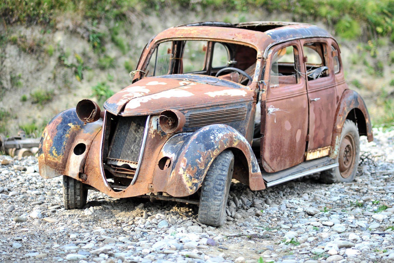 Rusty vintage car shell on rocky ground, with missing parts and faded red-brown paint.