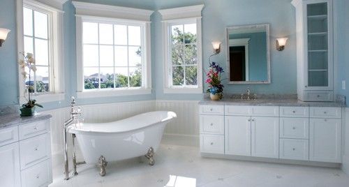 A bright bathroom with light blue walls, a claw-foot tub, white cabinetry, large windows, and a marble floor.