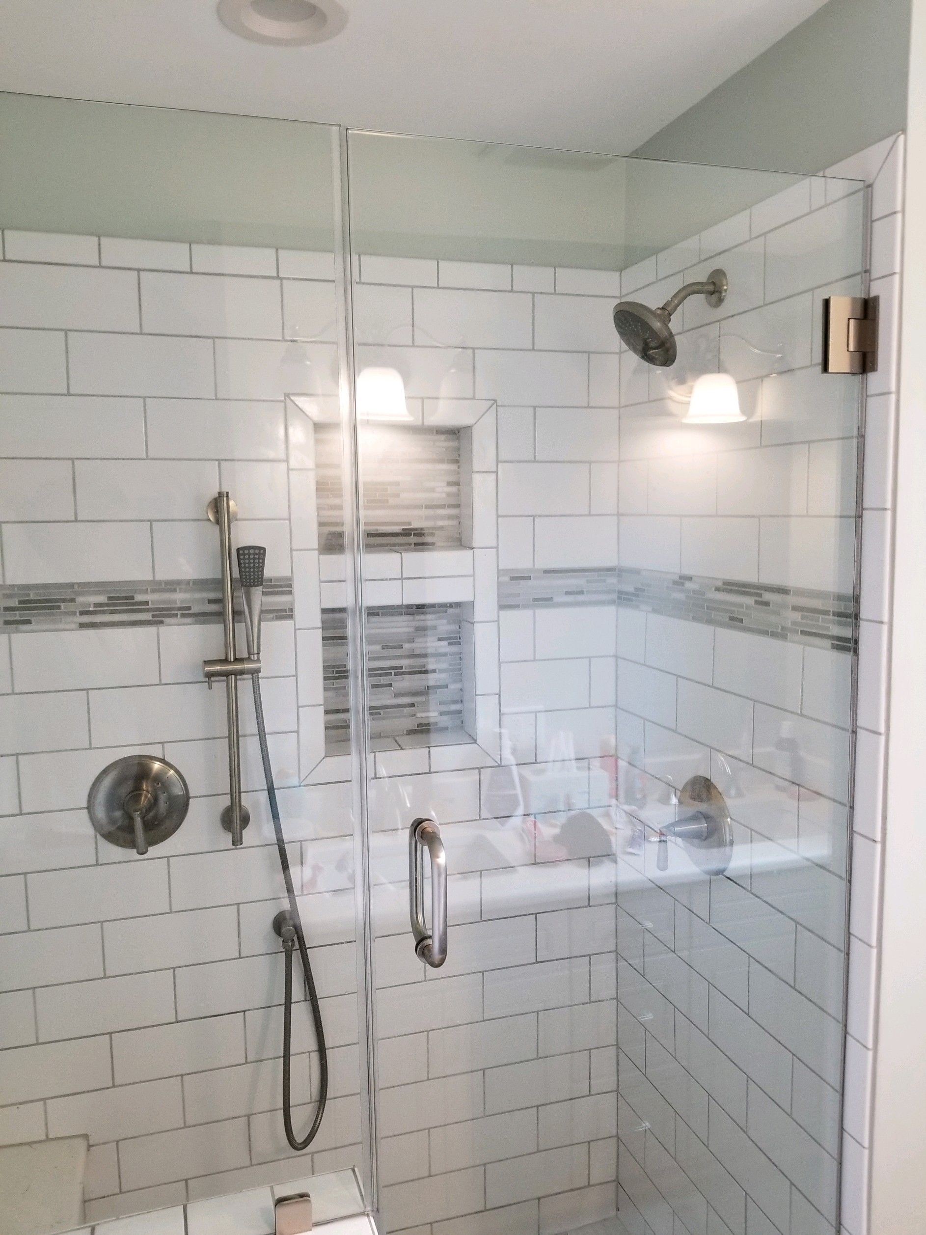 A walk-in shower with white subway tiles, a gray accent strip, two built-in niches, a rainfall showerhead, and glass doors.