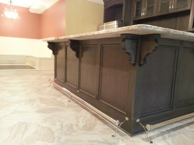 A dark wooden home bar with a marble countertop, decorative corbels, and a metal footrest, set on a light marble floor.