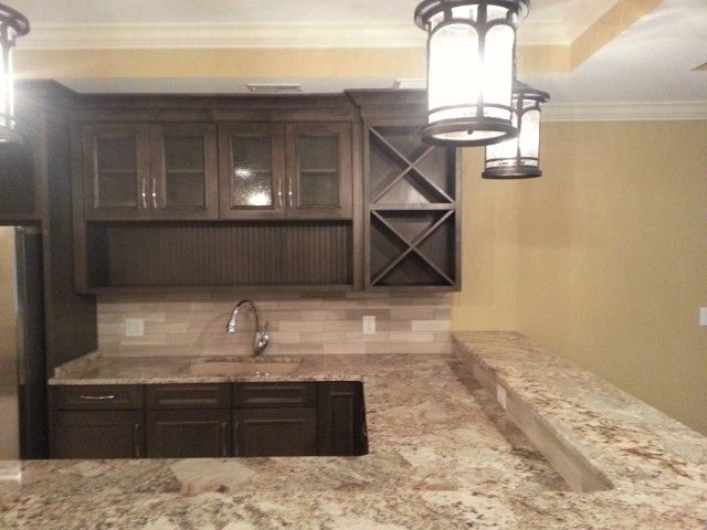 A dark wood kitchen bar with a granite countertop, a stainless steel sink, glass-front cabinets, and wine rack shelving.