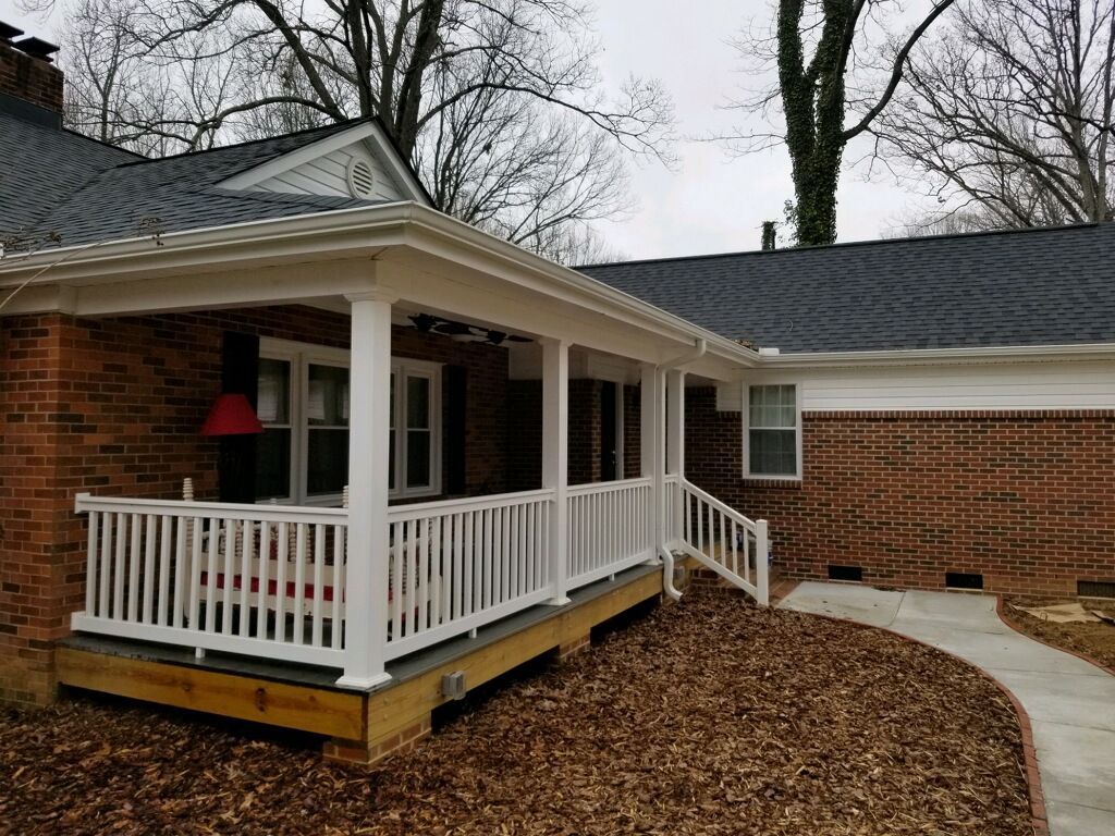 A new wooden porch with white railings and columns attached to a brick house with a dark roof and a concrete walkway.