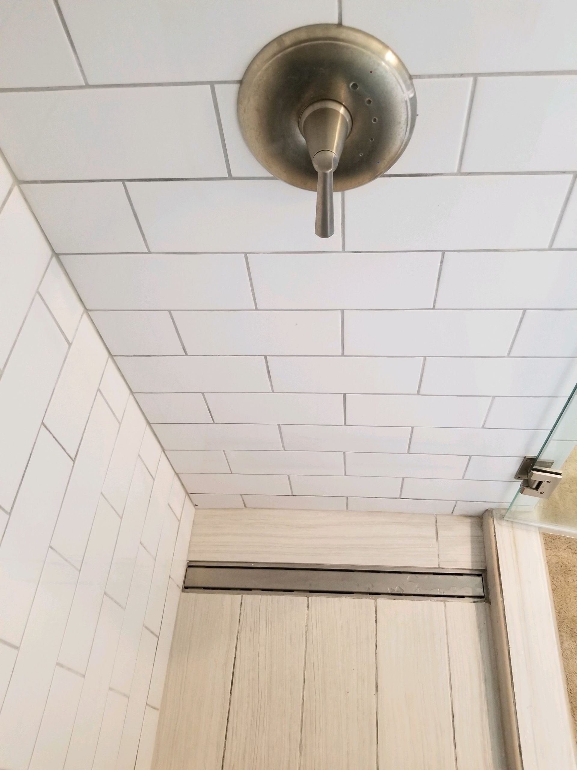 A shower interior with white rectangular wall tiles, a metallic control handle, and a linear floor drain near the entrance.