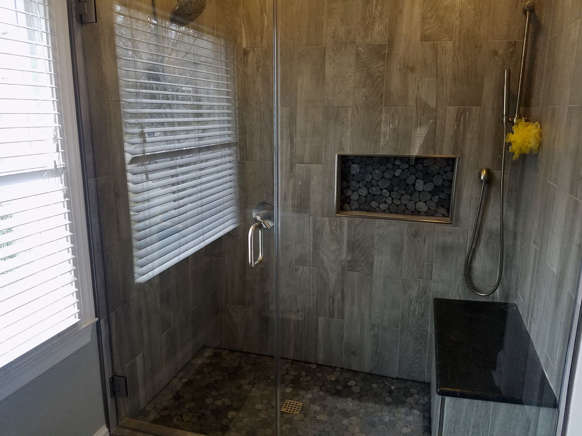 A modern walk-in shower with grey tile walls, a glass door, a black stone bench, and a rectangular tiled niche.
