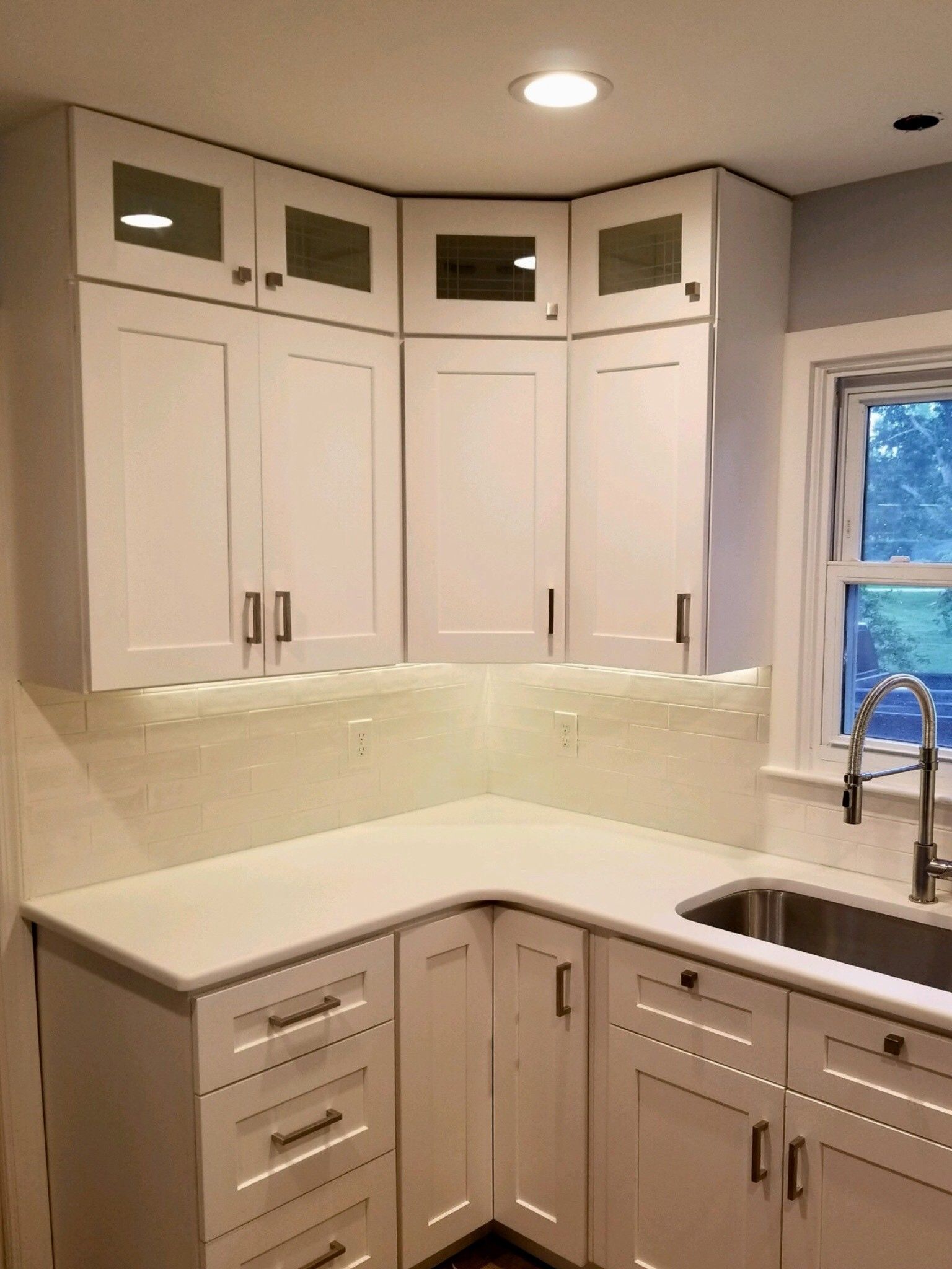 A white kitchen corner with cabinets, a matching countertop, a tile backsplash, and a sink by a window.