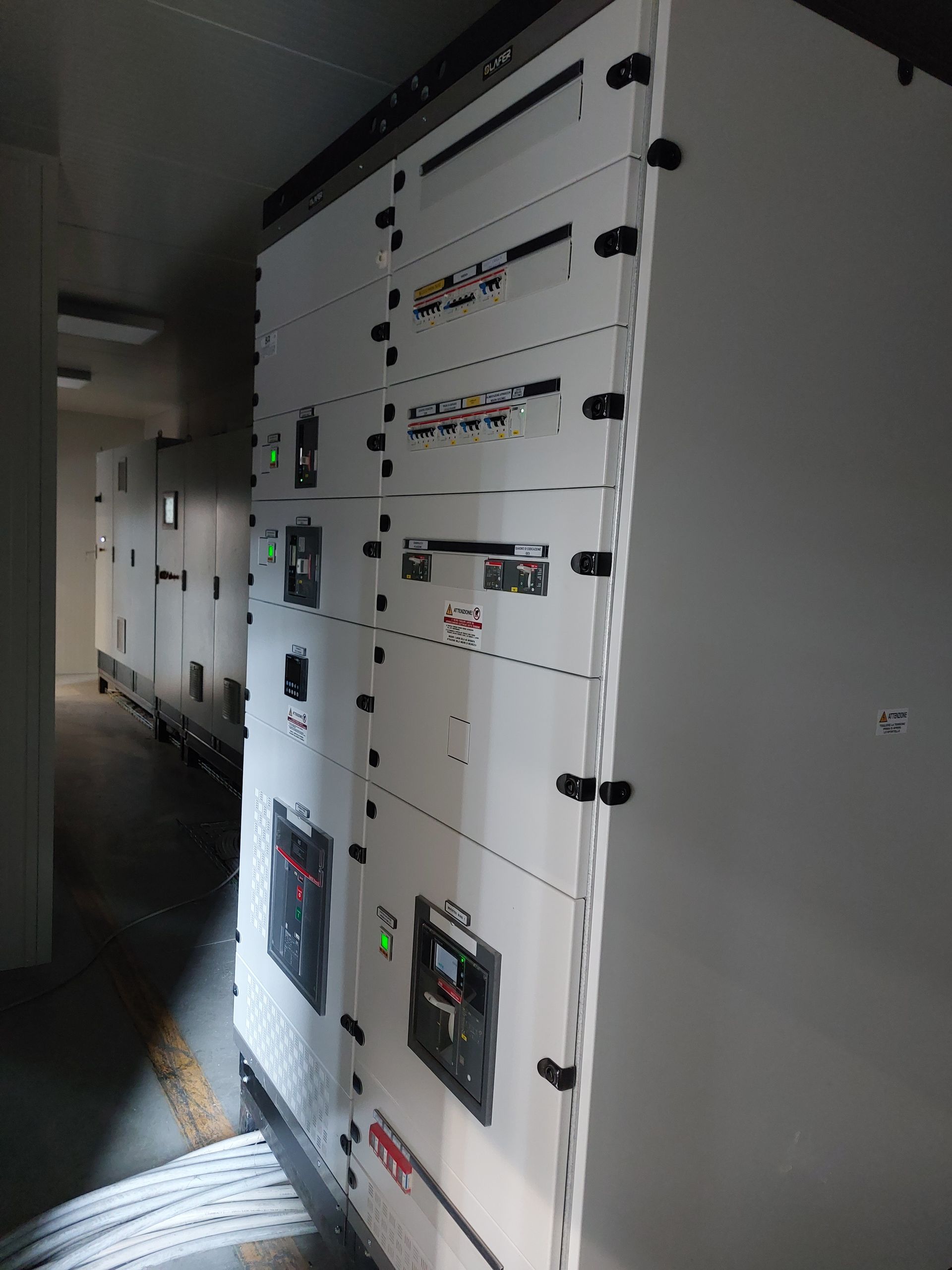 Quadri elettrici di controllo in un corridoio poco illuminato, con armadietti bianchi ed etichette di avvertenza.