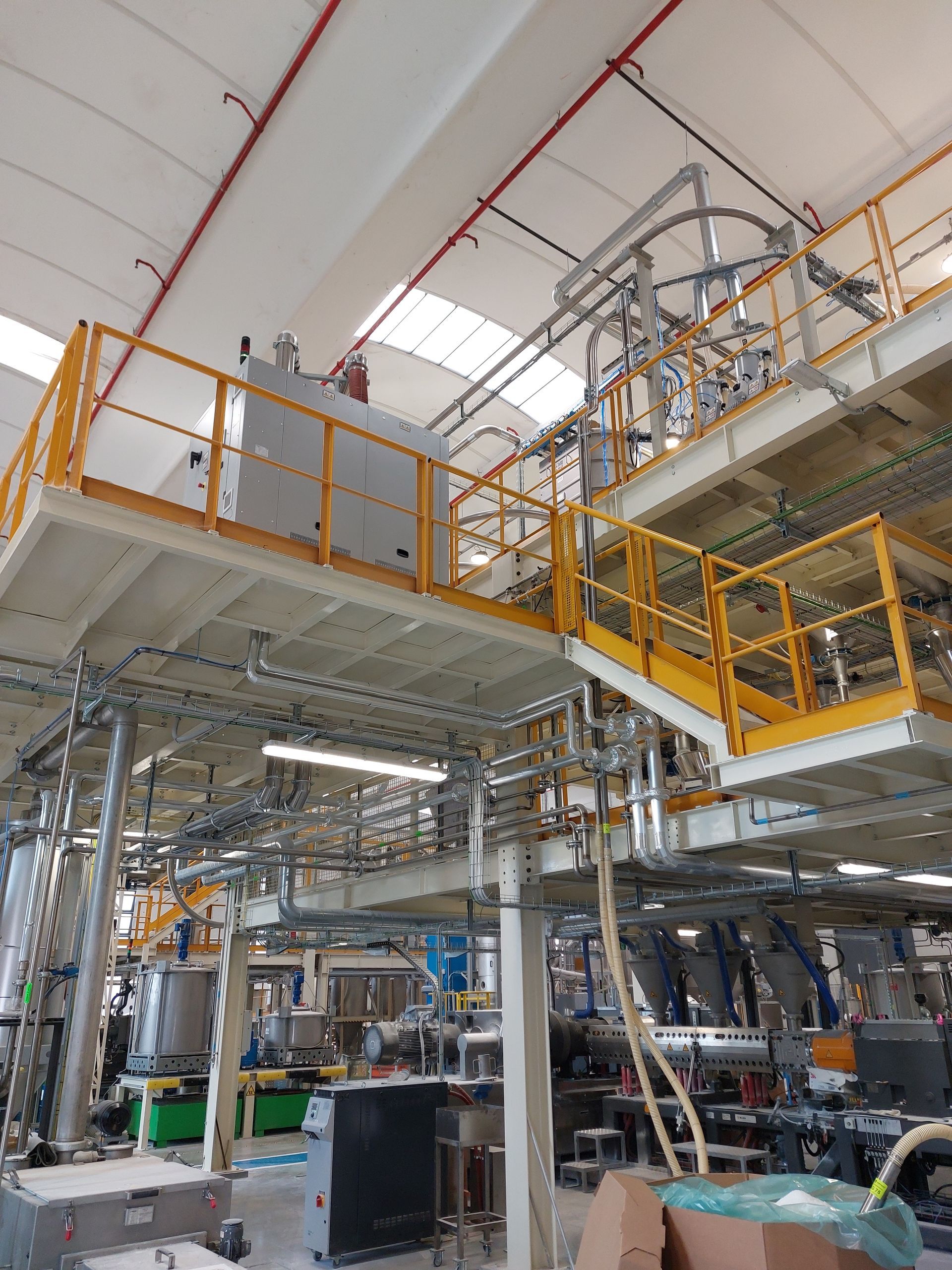 Interno di una fabbrica industriale con passerelle arancioni, tubature metalliche e macchinari sotto un alto soffitto bianco.