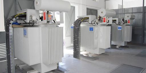 Trasformatori elettrici industriali bianchi in un locale simile a un magazzino, con tubi e cavi metallici.