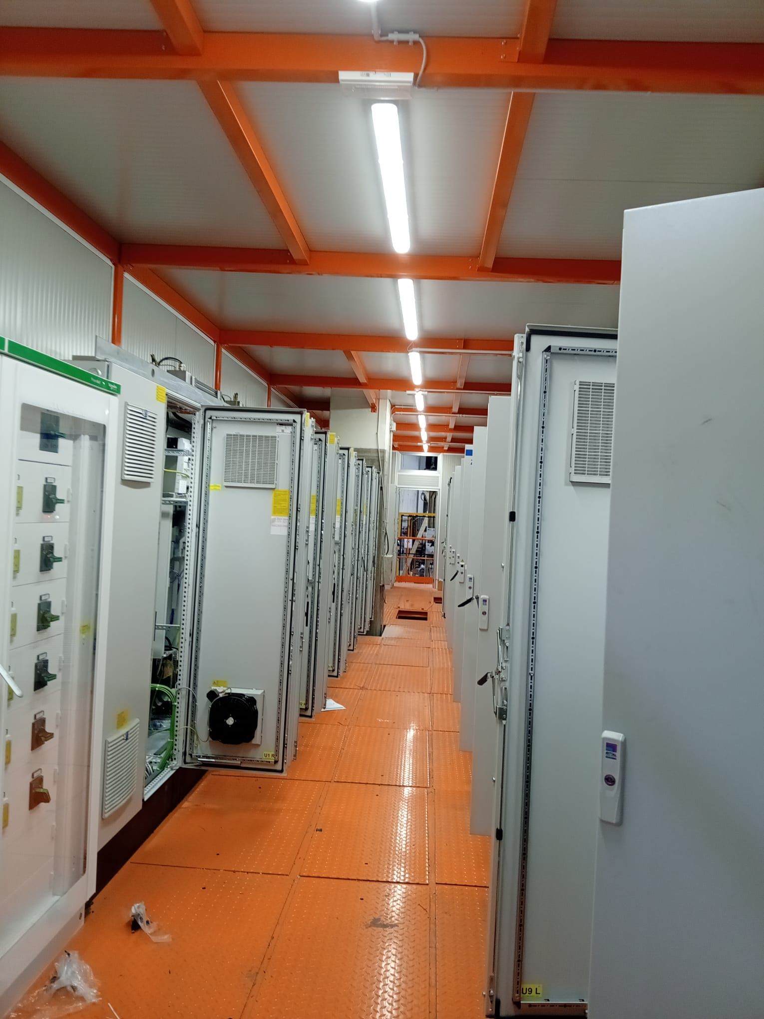 Uno stretto corridoio industriale con pavimento arancione, armadietti bianchi e travi a soffitto arancioni illuminate da luci fluorescenti.