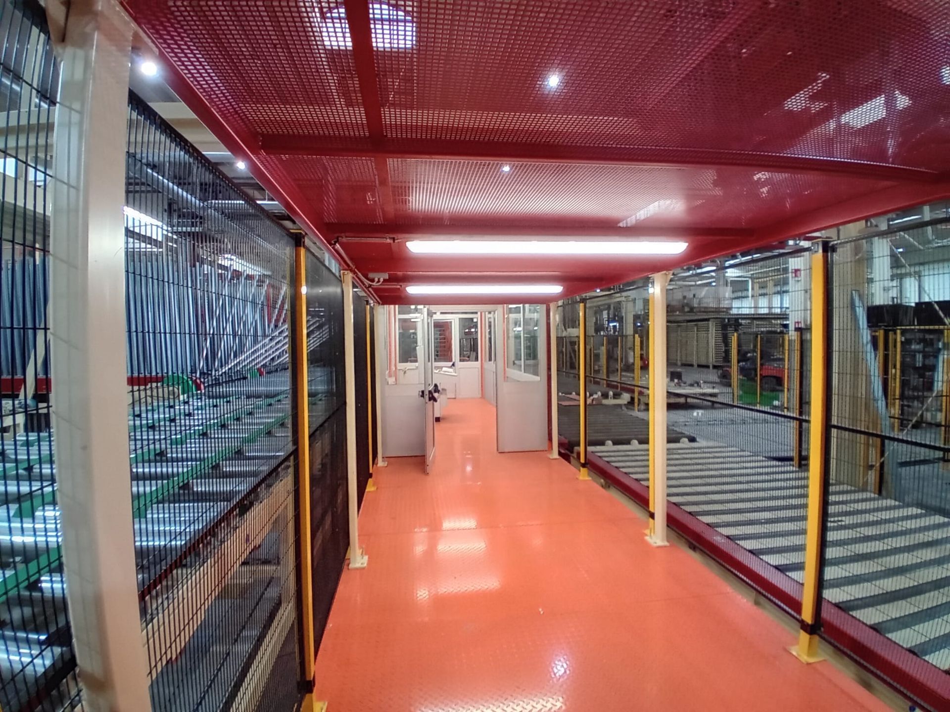 Corridoio industriale con pavimento e soffitto rossi, delimitato da recinzioni metalliche di sicurezza e macchinari su entrambi i lati.