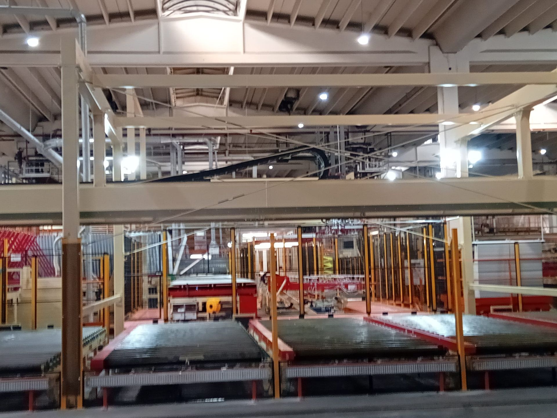 Interno di una fabbrica industriale con linee di trasporto, struttura metallica e travi a vista illuminate da luci intense.