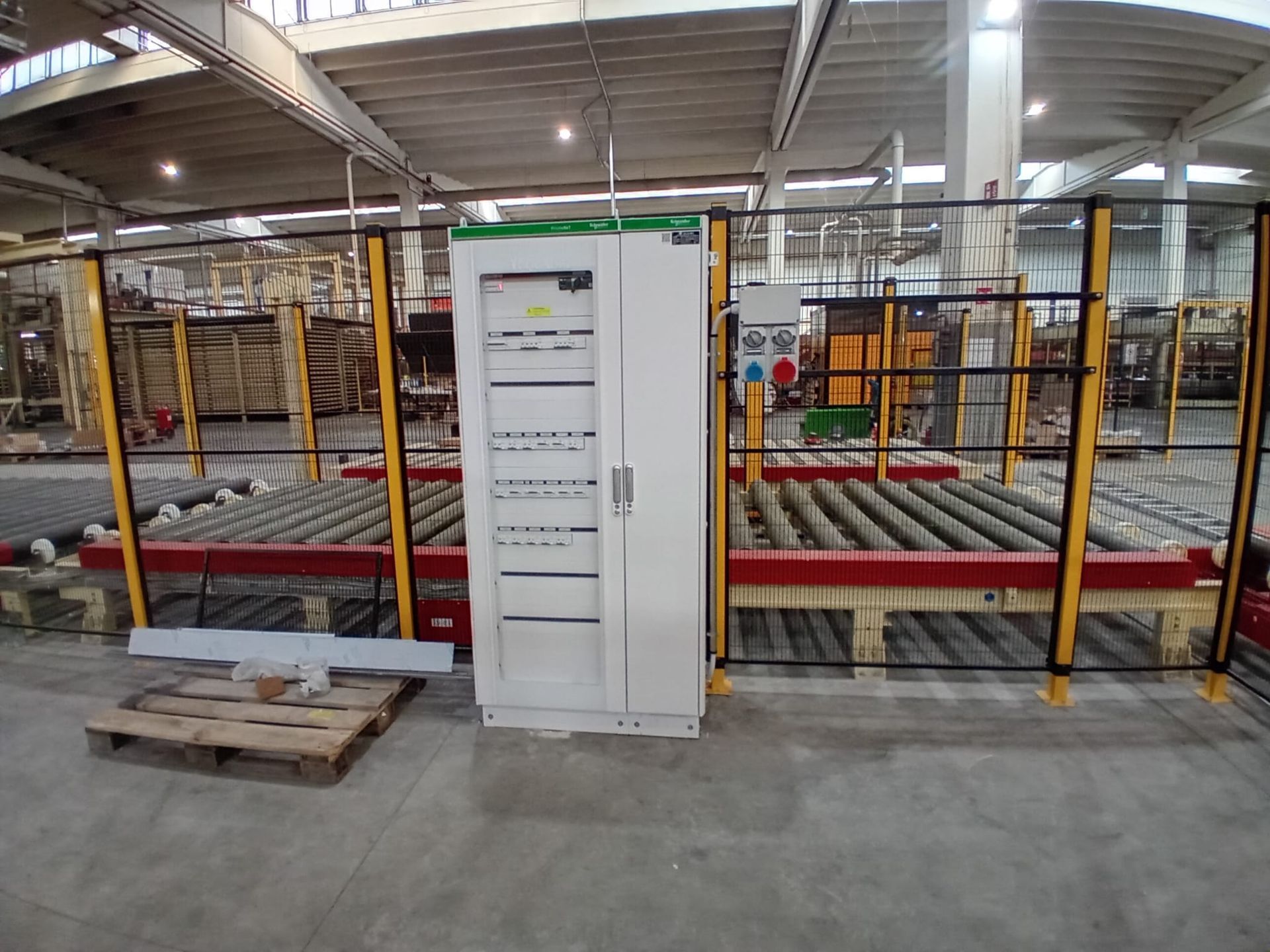 Magazzino industriale con rulli trasportatori, recinzione di sicurezza e un quadro elettrico bianco al centro.