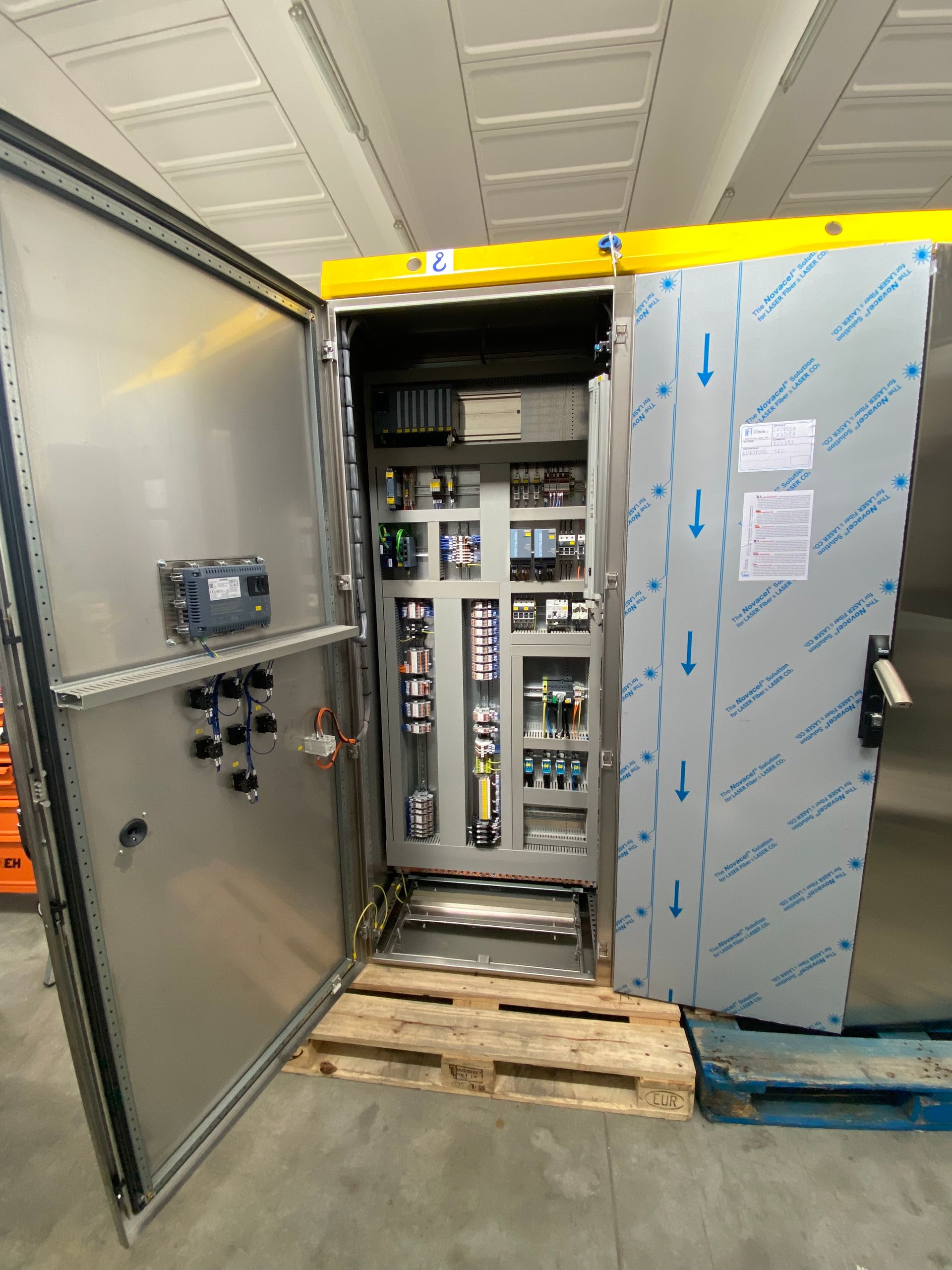 Armadio elettrico aperto con cablaggi e pannelli di controllo all'interno di un corridoio di magazzino.