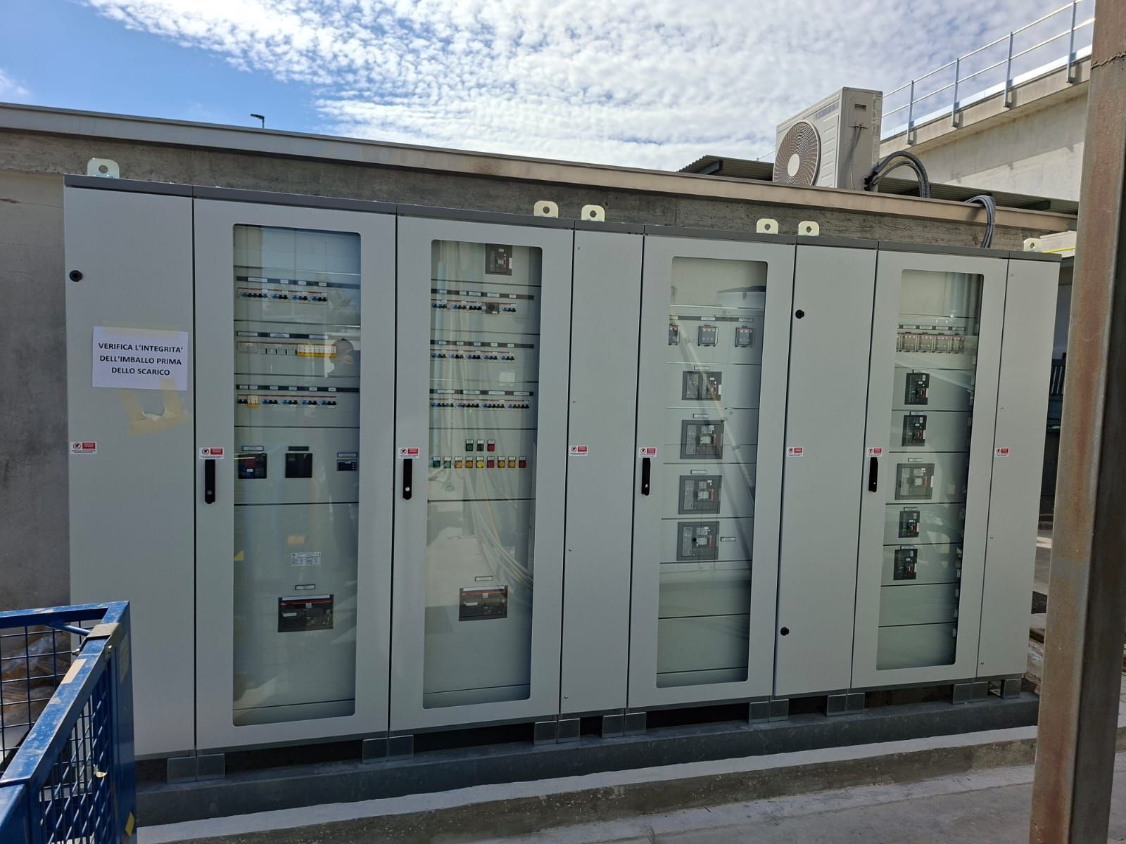 All'aperto, una fila di armadi elettrici grigi con ante in vetro ed etichette di avvertenza, accanto a un edificio sotto un cielo azzurro.