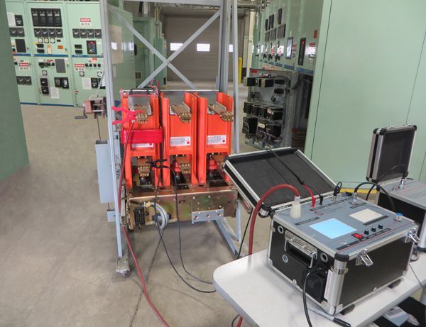 Apparecchiature elettriche in una sala di controllo con dispositivi arancioni su un carrello con ruote e uno strumento di prova su un tavolo.