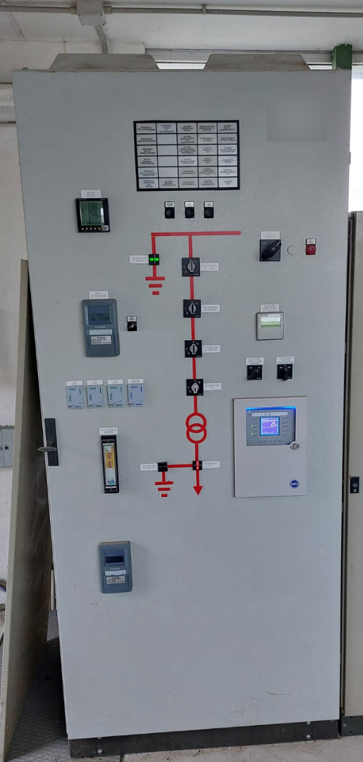 Pannello di controllo industriale grigio con interruttori, indicatori e un piccolo display in un locale tecnico.