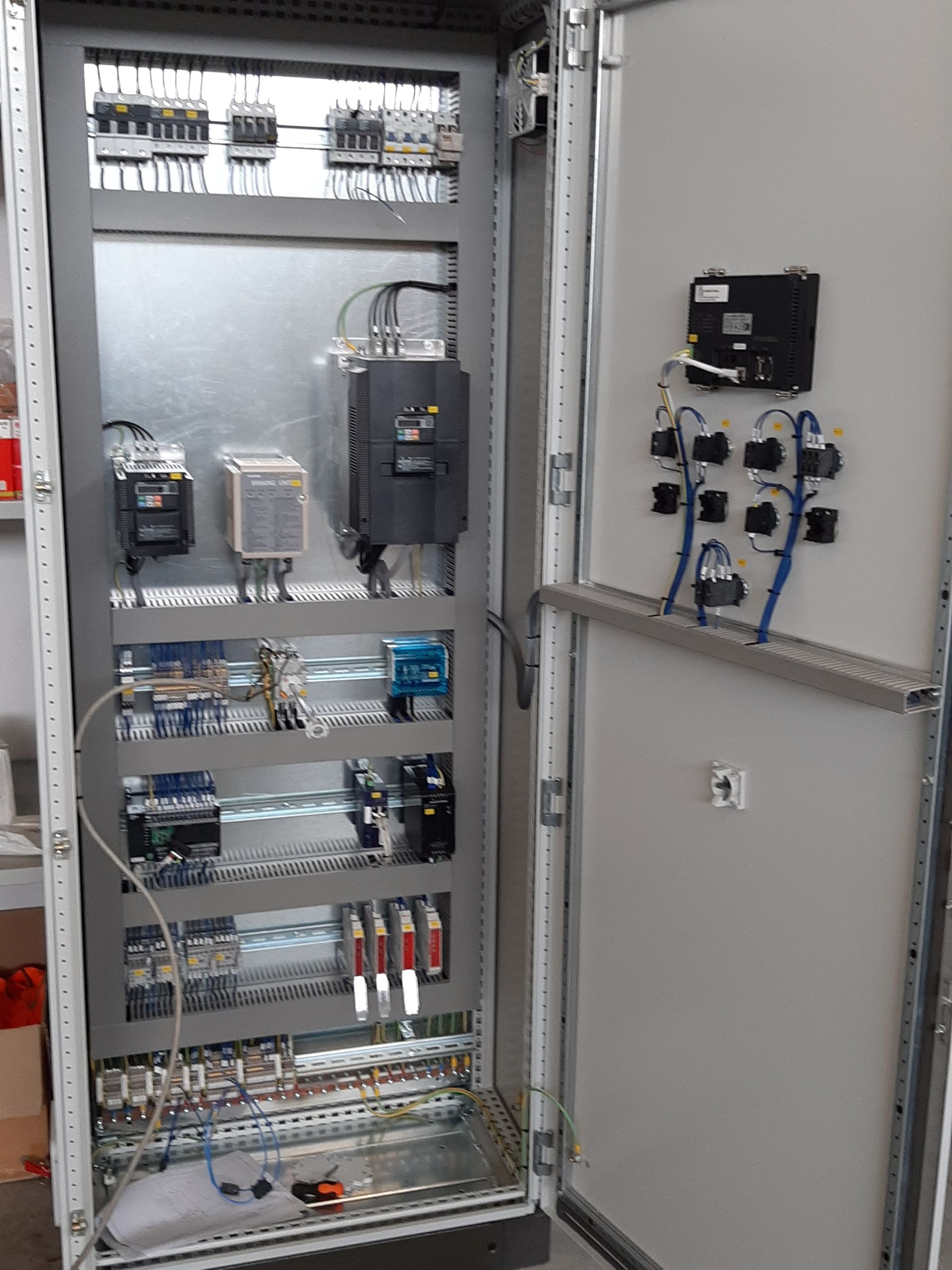 Quadro elettrico aperto con cablaggi, relè e componenti; l'anta dell'armadio contiene interruttori e indicatori.