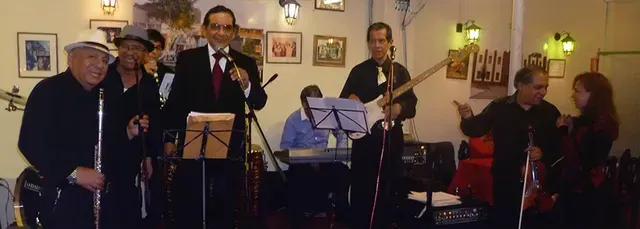 Músicos actuando en una sala con iluminación cálida, con un cantante, guitarristas y atriles para partituras.
