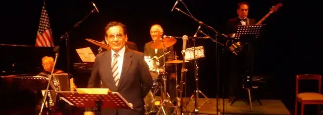 Un hombre habla desde un podio en un escenario oscuro, con un baterista y músicos detrás de él.