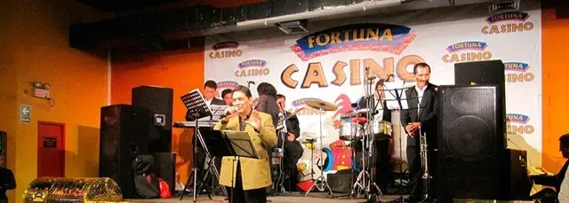 Grupo musical en directo actuando en un escenario iluminado con luces naranjas, con batería, micrófonos y un letrero de casino como telón de fondo.