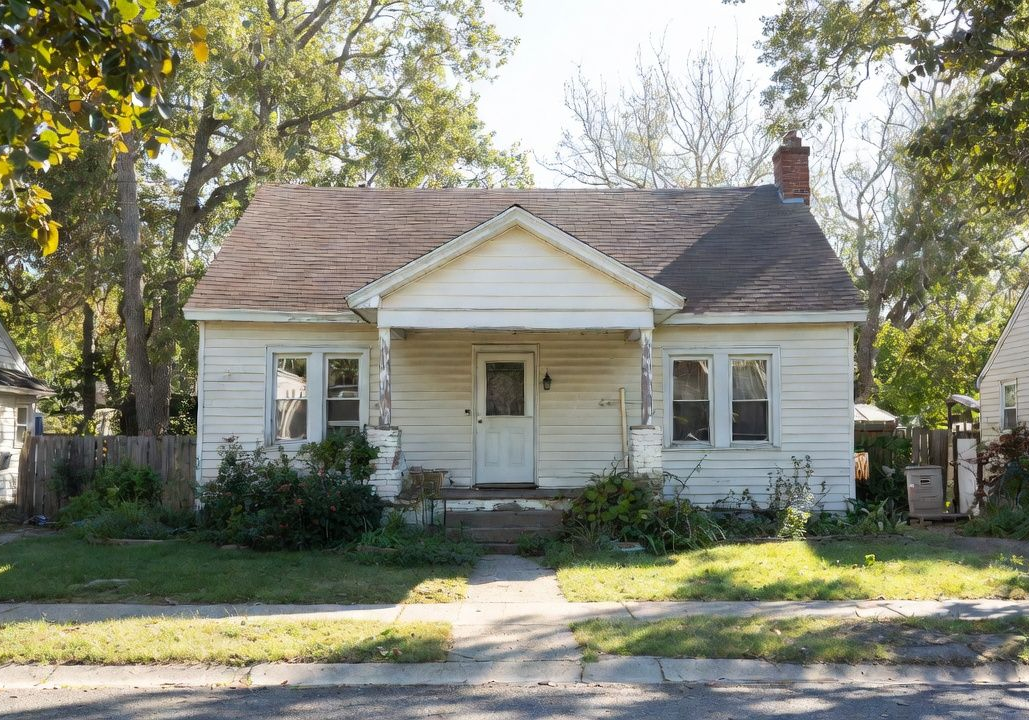 Small white house with a front porch, dark roof, and trees lining the street.